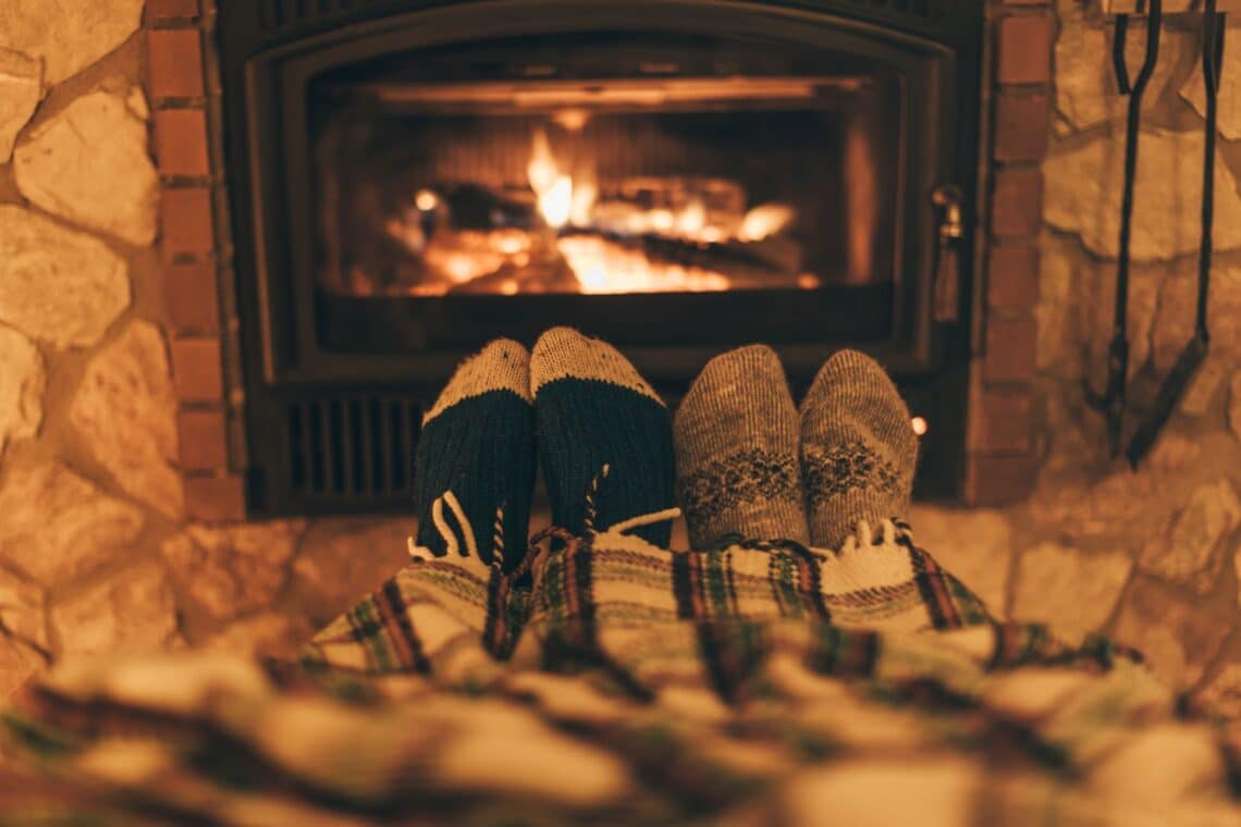 Zwei Personen, deren Füße in warmen Wollsocken vor einem lodernden Kaminfeuer unter einer karierten Decke hervorschauen. Der Kamin ist von einer Steinwand umgeben, die eine rustikale Atmosphäre schafft. Die warme Beleuchtung und das flackernde Feuer verstärken das Gefühl von Behaglichkeit und Entspannung.