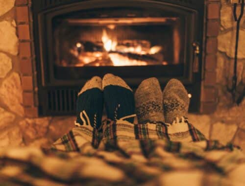 Zwei Personen, deren Füße in warmen Wollsocken vor einem lodernden Kaminfeuer unter einer karierten Decke hervorschauen. Der Kamin ist von einer Steinwand umgeben, die eine rustikale Atmosphäre schafft. Die warme Beleuchtung und das flackernde Feuer verstärken das Gefühl von Behaglichkeit und Entspannung.