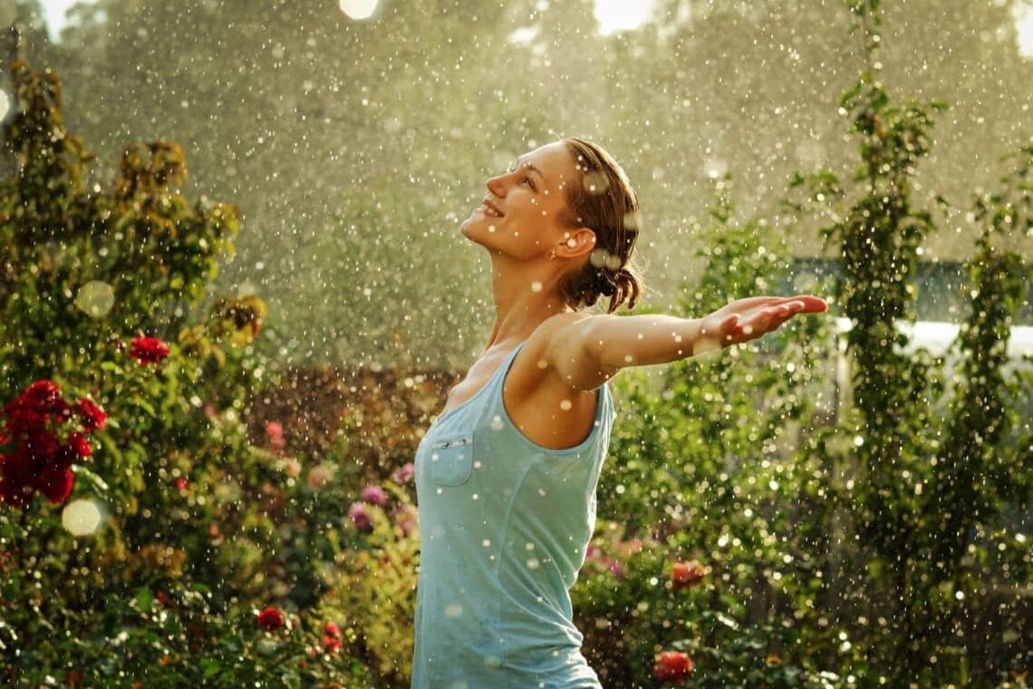 Eine Frau mit ausgestreckten Armen genießt den Regen im Garten, umgeben von Blumen und Bäumen, unter dem Schutz eines Pavillons in der Nähe.