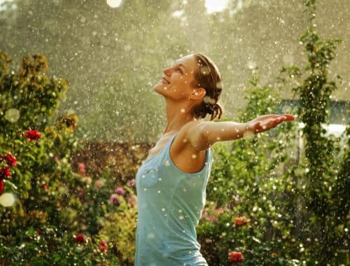 Eine Frau mit ausgestreckten Armen genießt den Regen im Garten, umgeben von Blumen und Bäumen, unter dem Schutz eines Pavillons in der Nähe.