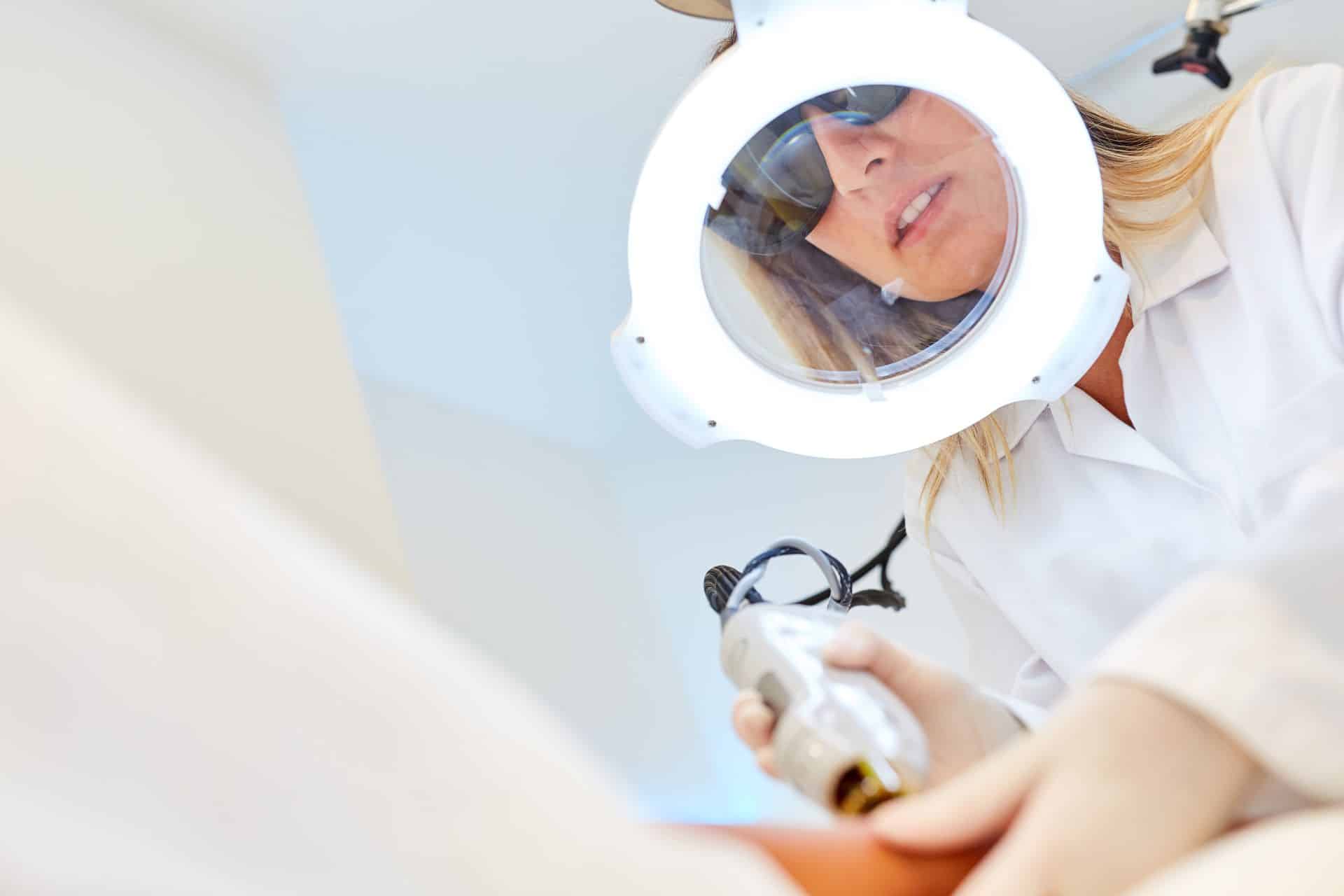 Kosmetikerin mit Schutzbrille und Lupenlampe führt eine Laser Haarentfernung an einer Kundin durch.