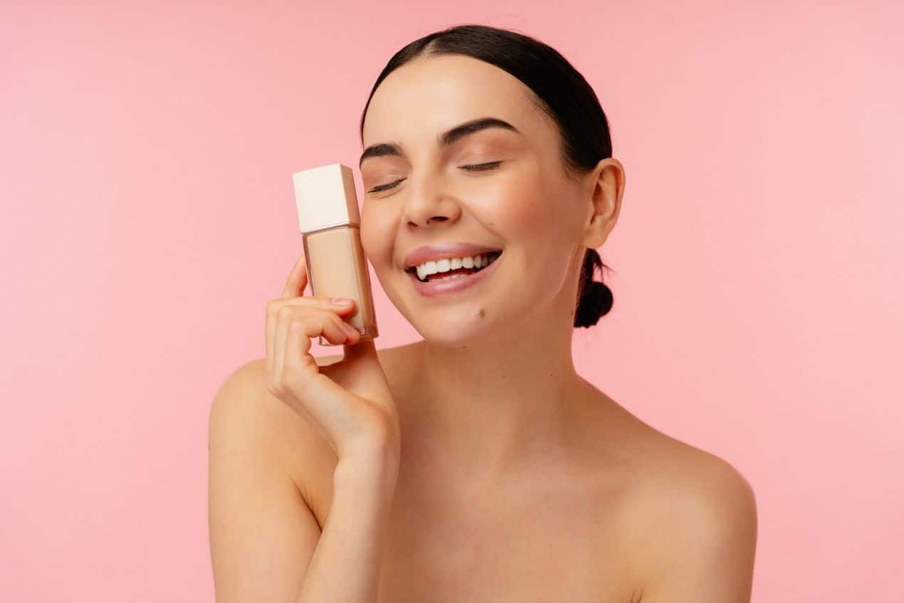 frau haelt jane iredale foundation flakon vor rosa hintergrund und laechelt