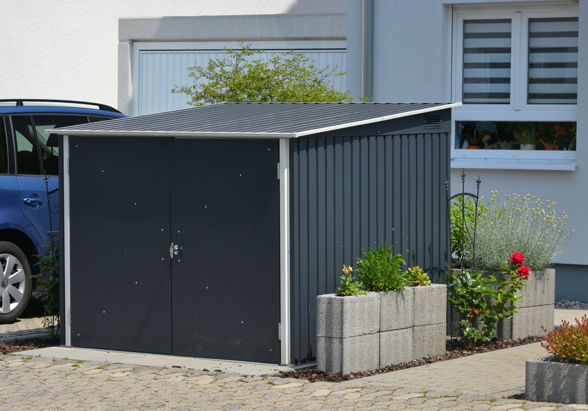 Abschliessbare Fahrradgarage aus Metall vor einem Wohnhaus mit Betonpflanzkübeln – wetterfest und platzsparend im Vorgarten platziert.